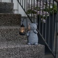 Solarna figurka ogrodowa LED w kształcie kota trzymającego latarenkę na haczyku, ustawiona na schodach przy balustradzie. Dekoracja solarna do ogrodu, na taras lub przy wejściu do domu, z podświetlaną latarnią tworzącą efekt świetlny.