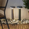 Dwie solarne lampy ogrodowe LED w kolorze beżowym i zielonym w formie słupków z panelem słonecznym, zamontowane wśród kamieni przy jasnej elewacji. Oświetlenie solarne do ogrodu, przy ścieżkach, rabatach lub tarasie.
