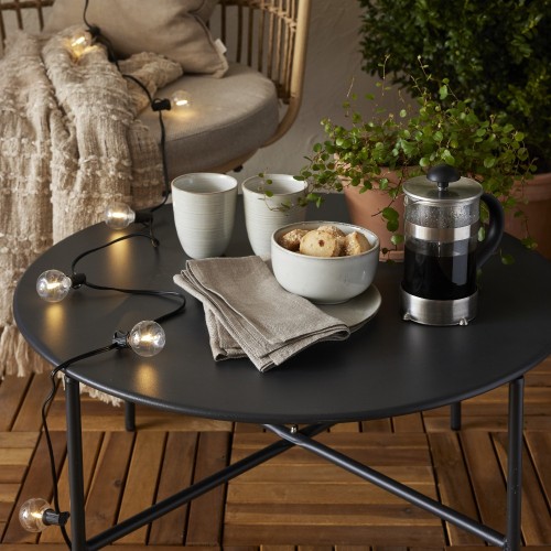 Solarna girlanda świetlna LED z dekoracyjnymi żarówkami w ciepłej barwie ułożona na czarnym stoliku tarasowym obok filiżanek, ciastek i zaparzacza do kawy. Oświetlenie solarne do ogrodu, na balkon lub taras, tworzące przytulny klimat.