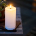 Biała świeca solarna LED w formie walca z realistycznym płomieniem ustawiona na drewnianym stoliku tarasowym. Dekoracyjna świeca solarna do ogrodu, na balkon lub taras.