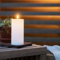 Biała świeca solarna LED w formie walca z realistycznym płomieniem ustawiona na drewnianym stoliku obok doniczki z rośliną. Dekoracyjna świeca solarna do ogrodu, na taras lub balkon.