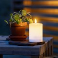 Biała świeca solarna LED w formie walca z realistycznym płomieniem ustawiona na drewnianym stoliku obok doniczki z rośliną. Dekoracyjna świeca solarna do ogrodu, na taras lub balkon.