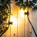 Girlanda ogrodowa z żarówkami LED E27 świecącymi ciepłym światłem na tle drewnianej ściany i dekoracji roślinnych.