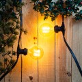 Zewnętrzna girlanda LED z żarówkami świecącymi ciepłym światłem zamocowana na drewnianej ścianie z dekoracjami roślinnymi. Nastrojowe oświetlenie do ogrodu i na taras.