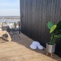 Solarne lampy ogrodowe LED w kształcie kamieni ustawione na drewnianym tarasie przy ścianie domu obok dużej donicy z rośliną. Dekoracyjne oświetlenie solarne na taras, balkon lub patio.
