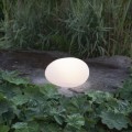 Lampa solarna LED w kształcie kamienia świecąca ciepłym światłem ustawiona na kamiennej płycie wśród roślin ogrodowych. Dekoracyjne oświetlenie solarne do ogrodu, na rabaty, ścieżki i taras.