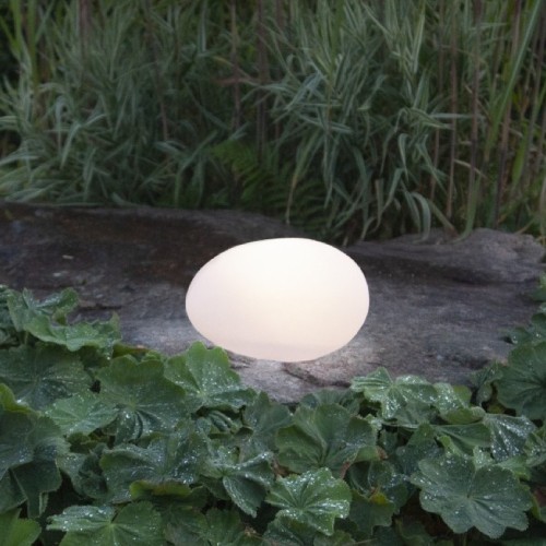 Lampa solarna LED w kształcie kamienia świecąca ciepłym światłem ustawiona na kamiennej płycie wśród roślin ogrodowych. Dekoracyjne oświetlenie solarne do ogrodu, na rabaty, ścieżki i taras.