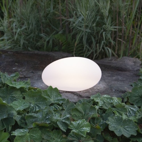 Solarna dekoracja ogrodowa LED w kształcie kamienia emitująca ciepłe światło, ustawiona na kamiennej płycie wśród roślin. Dekoracyjne oświetlenie solarne do ogrodu, na rabaty, ścieżki i taras.