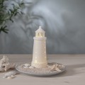 Biała porcelanowa latarnia morska LED na dekoracyjnej podstawie z piaskiem i muszlami ustawiona na stole jako element aranżacji wnętrza w stylu nadmorskim emitująca ciepłe światło.