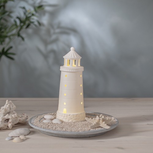 Biała porcelanowa latarnia morska LED na dekoracyjnej podstawie z piaskiem i muszlami ustawiona na stole jako element aranżacji wnętrza w stylu nadmorskim emitująca ciepłe światło.