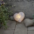 Dekoracyjne serce LED z efektem kamienia emitujące ciepłe światło, umieszczone przy nagrobku wśród roślin i kamieni, jako subtelna dekoracja pamięci.