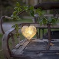 Dekoracyjne serce LED z efektem kamienia ustawione na drewnianym krześle w ogrodzie, emitujące ciepłe światło, otoczone zielonymi liśćmi, idealne jako nastrojowa ozdoba do przestrzeni zewnętrznych.