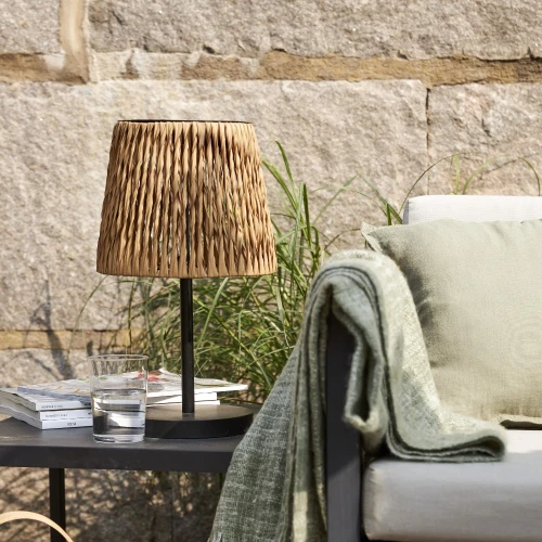 Solarna lampa stołowa ogrodowa LED z plecionym abażurem na stoliku przy sofie, obok książek i szklanki wody, dekoracyjne oświetlenie tarasu.