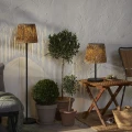 Solarna lampa podłogowa i stołowa ogrodowa LED z plecionym abażurem przy roślinach doniczkowych i drewnianym stoliku, ciepłe światło dekoracyjne.