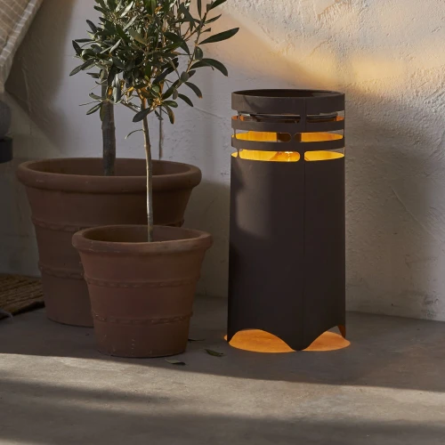 Solarna lampa ogrodowa LED z funkcją ładowania USB, emitująca ciepłe światło, ustawiona obok donic z roślinami przy ścianie.