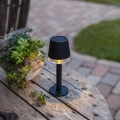 Solarna lampa stołowa LED z panelem na górze, ustawiona na drewnianym stole obok roślin.