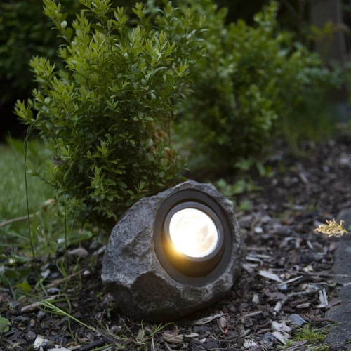 Solarny reflektor ogrodowy LED w formie kamienia ustawiony przy ścieżce wśród roślin, delikatne oświetlenie rabaty i przestrzeni ogrodowej.