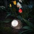 Solarny reflektor ogrodowy LED w formie kamienia umieszczony wśród kwiatów, oświetla rośliny i rabatę delikatnym światłem po zmroku.
