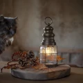 Stylowa lampa w kształcie latarni z ciepłym światłem, ustawiona na stole w towarzystwie naturalnych dekoracji, idealna do stworzenia nastrojowej aranżacji wnętrza.