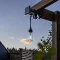 Solarna lampa ogrodowa w formie żarówki zawieszona na drewnianej konstrukcji, emitująca ciepłe światło i tworząca nastrojowy klimat o zmierzchu.