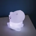 Lampka nocna w kształcie dinozaura świecąca delikatnym białym światłem, ustawiona na stoliku nocnym przy ścianie, idealna jako subtelne oświetlenie do sypialni.