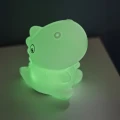 Lampka nocna dinozaur świecąca zielonym światłem, ustawiona na stoliku jako dekoracyjne i nastrojowe oświetlenie do pokoju.