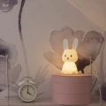 Lampka nocna w kształcie króliczka emitująca ciepłe światło, ustawiona na komodzie obok budzika w przytulnym pokoju dziecięcym.