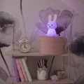Lampka nocna króliczek w fioletowym świetle, ustawiona na półce z książkami i dekoracjami w pokoju dziecięcym.