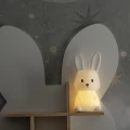 Lampka nocna w kształcie króliczka emitująca ciepłe światło, ustawiona na półce w pokoju dziecięcym jako przytulne i dekoracyjne oświetlenie.