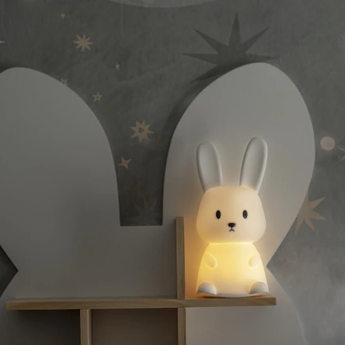 Lampka nocna w kształcie króliczka emitująca ciepłe światło, ustawiona na półce w pokoju dziecięcym jako przytulne i dekoracyjne oświetlenie.