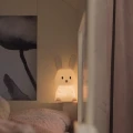 Lampka nocna króliczek świecąca ciepłym światłem, ustawiona przy łóżku jako spokojne oświetlenie nocne dla dziecka.