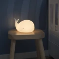 Lampka nocna w kształcie wieloryba ustawiona na drewnianym stołku przy łóżeczku, emitująca ciepłe, delikatne światło idealne do nocnego oświetlenia.