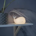 Lampka nocna w kształcie wieloryba stojąca na półce obok rośliny, emitująca ciepłe, delikatne światło w przytulnym wnętrzu.