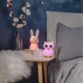 Lampki nocne w kształcie króliczka i sowy z delikatnym, różowym światłem, ustawione na stoliku przy łóżku, wprowadzają przytulny i spokojny nastrój do wnętrza.