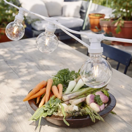 Girlanda świetlna z żarówkami LED G95 1W filament o ciepłej barwie 2700K na białym przewodzie, zawieszona nad stołem ogrodowym z warzywami. Dekoracyjne oświetlenie do ogrodu, tarasu i przestrzeni zewnętrznych.