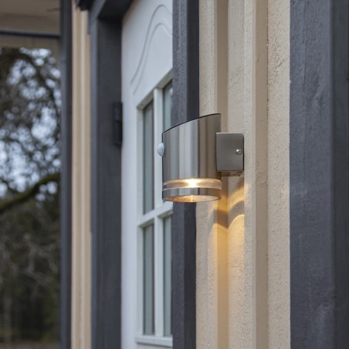 Kinkiet solarny z czujnikiem ruchu zamontowany przy drzwiach wejściowych, emituje ciepłe światło LED i subtelnie podkreśla elewację budynku.