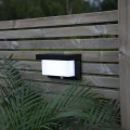 Kinkiet solarny na drewnianym płocie z białym panelem świetlnym, zapewnia funkcjonalne oświetlenie przestrzeni przy roślinach, podkreślając estetykę ogrodu po zmroku.