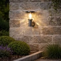 Solarny kinkiet ogrodowy z dekoracyjnym kloszem emituje ciepłą barwę światła, tworząc przytulny efekt na kamiennej elewacji i subtelnie podkreślając roślinność wokół.