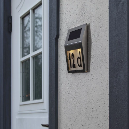 Solarna lampa z podświetlanym numerem domu „12d”, zamontowana na elewacji obok drzwi wejściowych.
