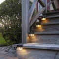 Cztery solarne lampy LED zamontowane na drewnianych schodach tarasowych emitują ciepłe światło, subtelnie oświetlając stopnie i zwiększając bezpieczeństwo po zmroku w ogrodzie lub przy domu.