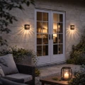 Dwa solarne kinkiety zamontowane po obu stronach drzwi tarasowych emitują ciepłe światło, tworząc dekoracyjny efekt cieni i przytulną atmosferę przy wejściu do domu po zmroku.