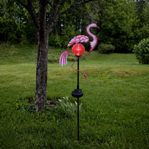 Solarna dekoracja ogrodowa flaming na szpikulcu z podświetlaną kulą, ustawiona przy trawniku i drzewie, tworzy przyjemny nastrój po zmroku.