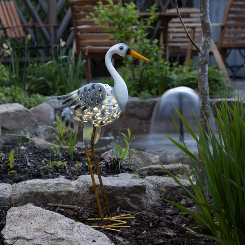 Zbliżenie na dekoracyjną czaplę LED z ażurowym korpusem i ciepłym światłem, ustawioną przy oczku wodnym, podkreśla jej elegancki detal i naturalne dopasowanie do ogrodu.