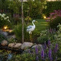 Metalowa figurka czapli z ażurowymi skrzydłami stoi przy kamiennej rabacie obok oczka wodnego, a subtelne podświetlenie LED wydobywa jej detale, tworząc spokojny, elegancki akcent w ogrodzie.