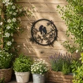 Czarna, okrągła dekoracja solarna z motywem dwóch ptaków na gałęzi, zamocowana na drewnianej ścianie. Wokół widoczne donice z kwitnącymi roślinami i zielenią, tworzące naturalne, przytulne otoczenie.