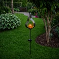 Solarna lampa ogrodowa na szpikulcu w kształcie płomienia z dekoracyjną, bursztynową kulą LED. Umieszczona na trawniku wśród roślin, tworzy wyrazisty punkt świetlny i podkreśla charakter zielonej przestrzeni.