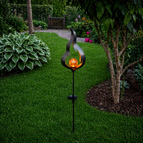 Solarna lampa ogrodowa na szpikulcu w kształcie płomienia z dekoracyjną, bursztynową kulą LED. Umieszczona na trawniku wśród roślin, tworzy wyrazisty punkt świetlny i podkreśla charakter zielonej przestrzeni.