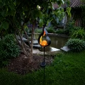 Solarna lampa ogrodowa w formie płomienia z bursztynową kulą LED, umieszczona przy drzewie obok oczka wodnego. Ciepłe światło tworzy przyjemny klimat i subtelnie podkreśla naturalne otoczenie ogrodu.