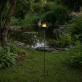 Solarna lampa ogrodowa w kształcie półksiężyca z kulą LED, ustawiona przy oczku wodnym. Ciepłe światło delikatnie odbija się w wodzie i podkreśla naturalny charakter zielonej przestrzeni.