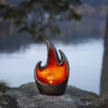 Solarna lampa LED z efektem płomienia i bursztynową kulą, ustawiona na kamieniu przy wodzie. Delikatne światło nadaje otoczeniu spokojny, naturalny charakter.